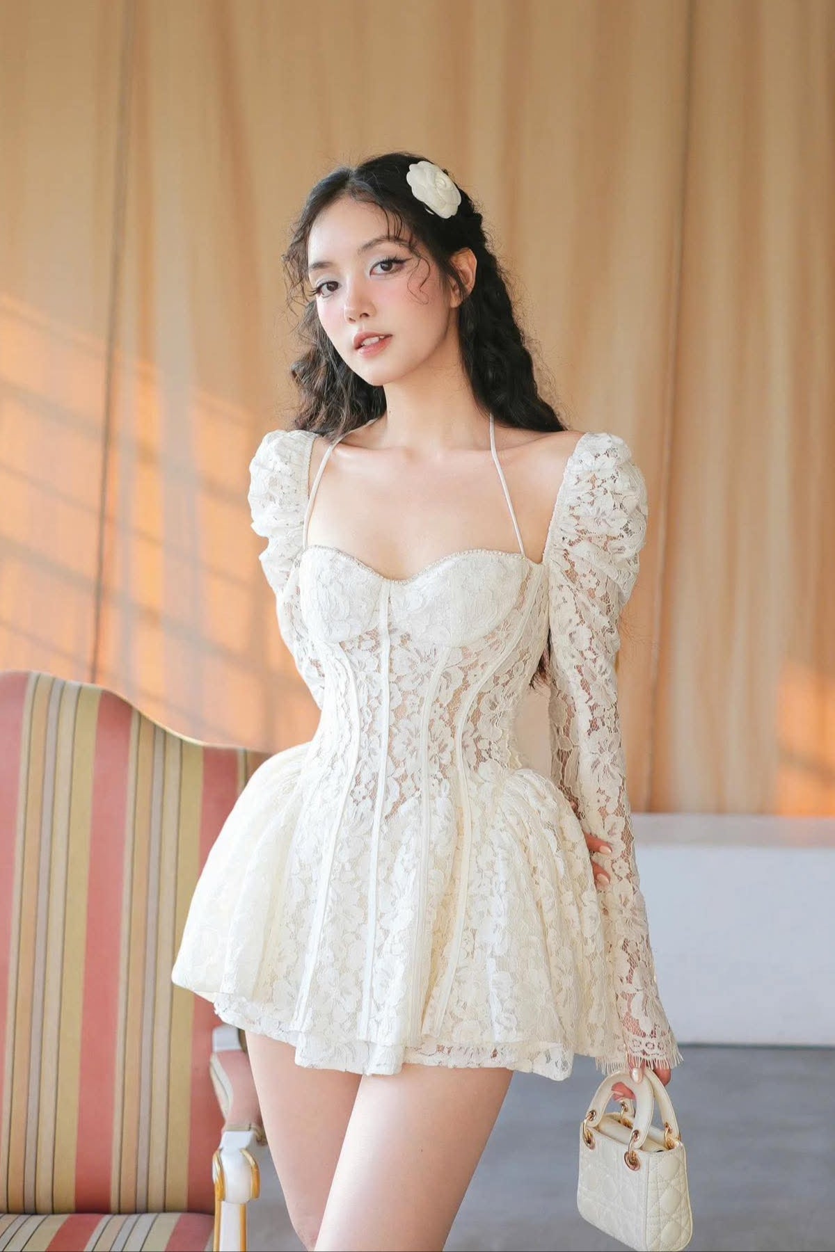 Woman in a white lace dress standing in a softly lit room with curtains.