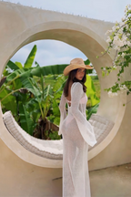 Woman in a white dress and straw hat standing in front of a circular concrete structure with greenery in the background.