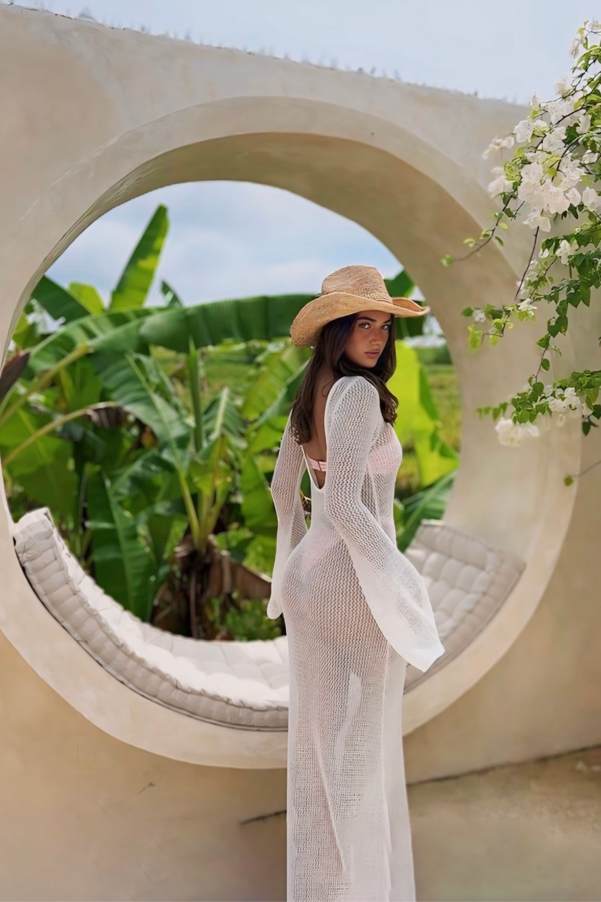Woman in a white dress and straw hat standing in front of a circular concrete structure with greenery in the background.