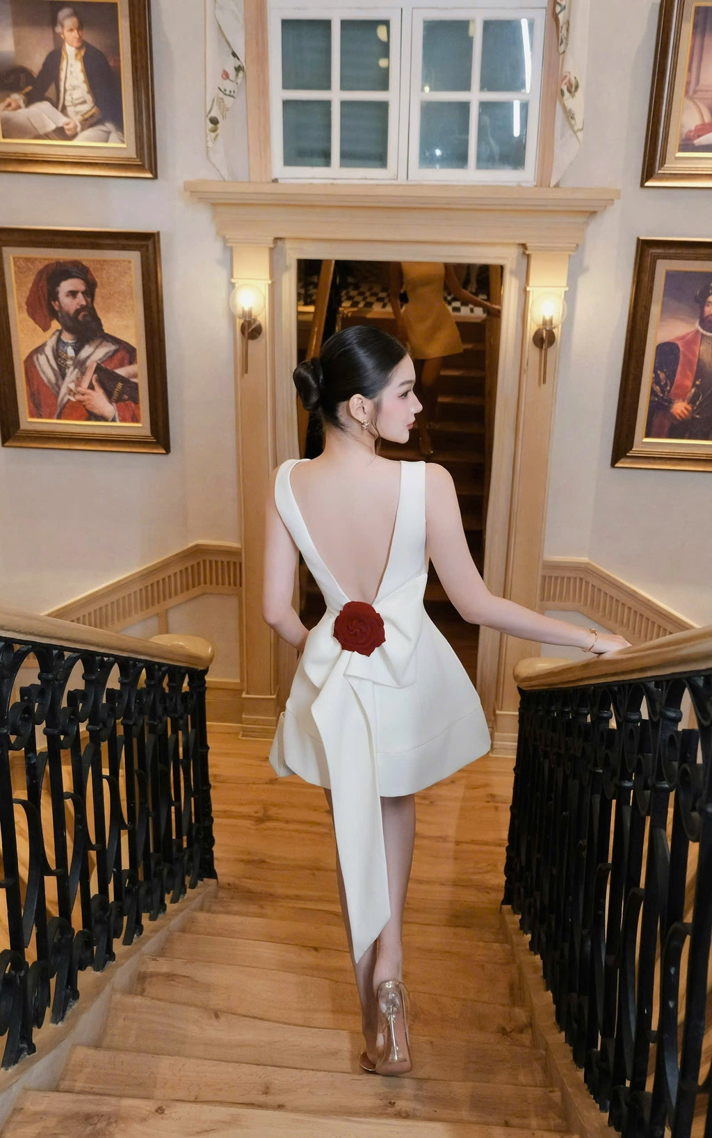 Woman in a white dress with a red flower walking down a staircase in an elegant interior setting.