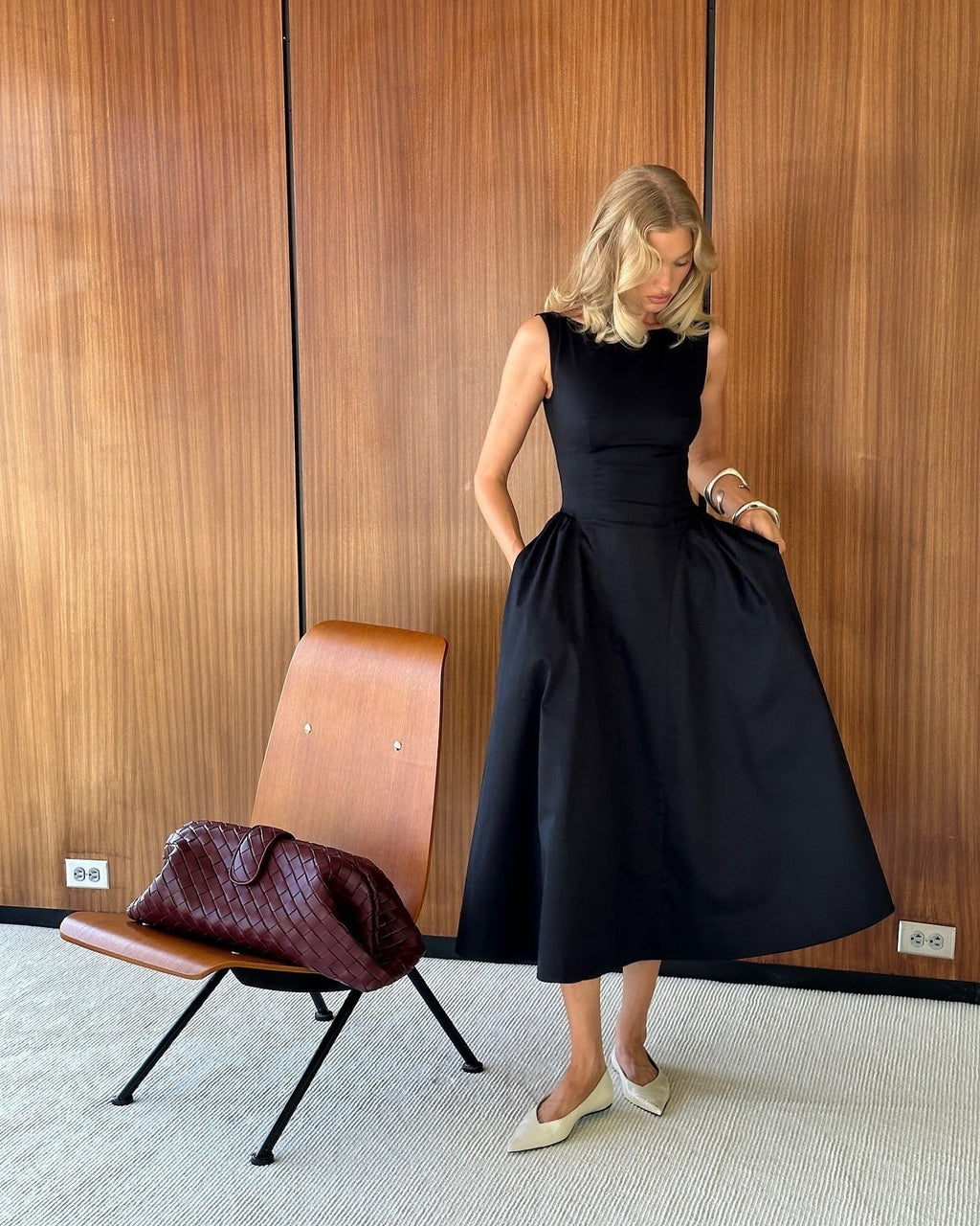 Woman in a black dress standing next to a brown chair in a room with wooden paneling.